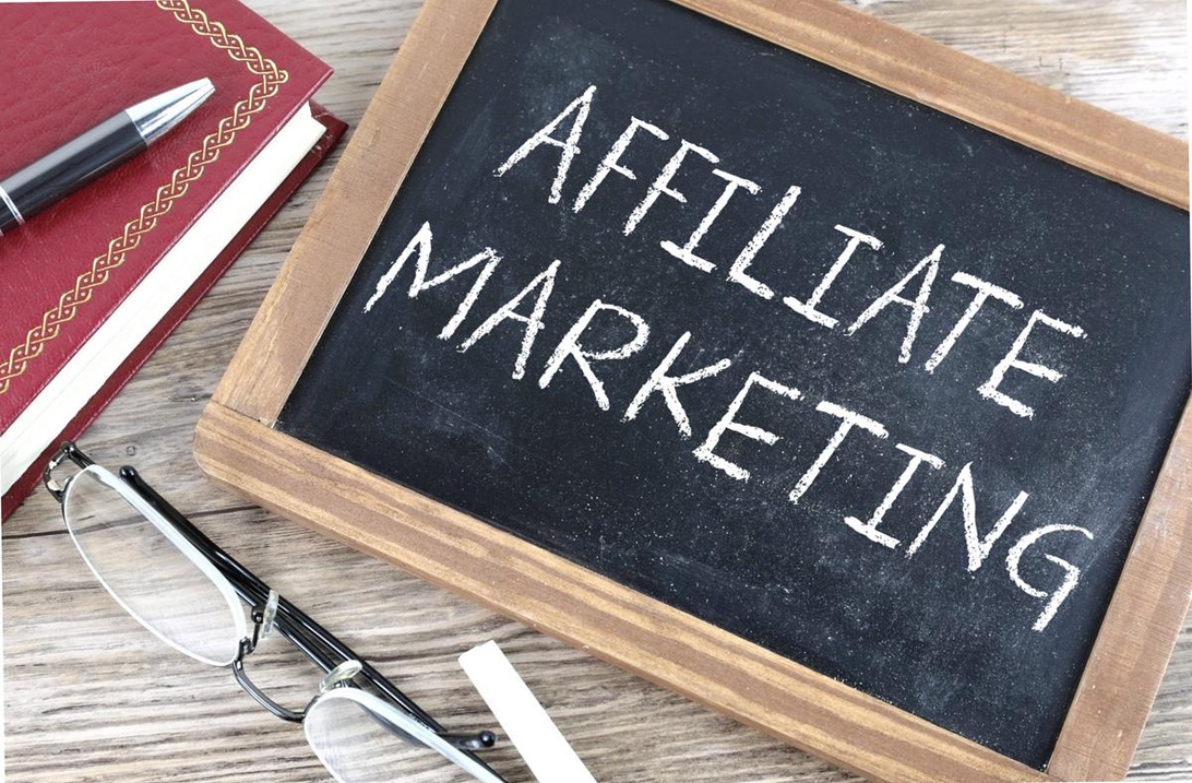 En lille sort tavle med 'AFFILIATE MARKETING' håndskrevet med kridt. Bøger, en kuglepen og læsebriller ligger i nærheden på en træoverflade.